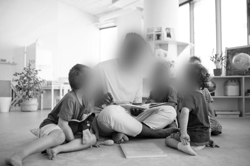 Un maestro se sienta en el suelo leyendo a cuatro niños pequeños en un aula luminosa. Los niños están reunidos alrededor, escuchando atentamente.