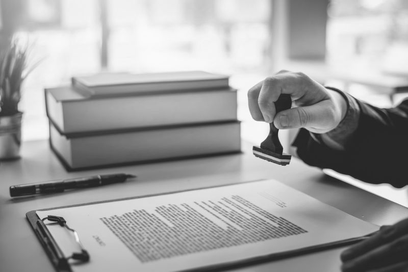 Eine Hand hält einen Stempel und drückt ihn auf ein Dokument auf einem Schreibtisch mit einem Stift und einem Bücherstapel. Die Szene ist schwarz-weiß.