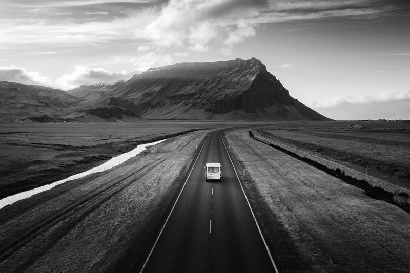 Fotografía en blanco y negro de gran angular, en el centro se encuentra un coche blanco, al fondo un paisaje austero: praderas y una cadena montañosa