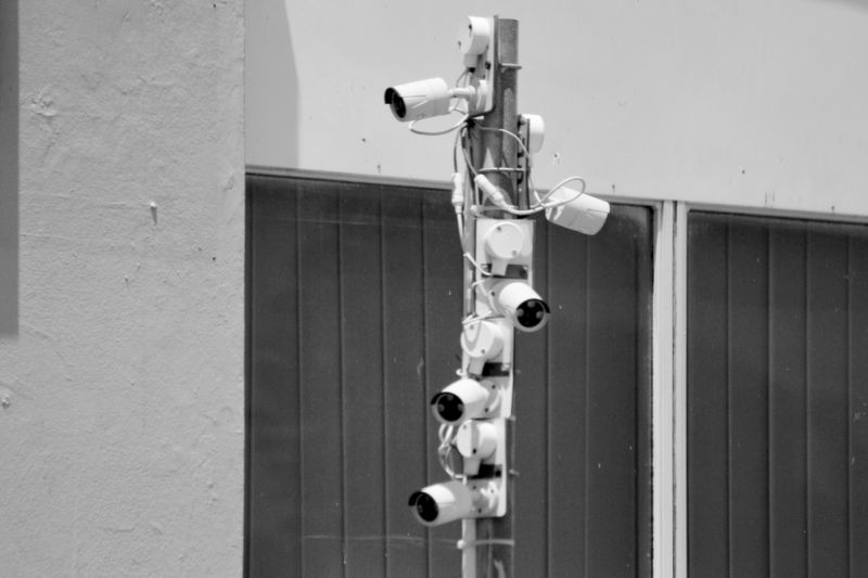 Un poste vertical con ocho cámaras de vigilancia instaladas, situado frente a una pared con grandes ventanas al fondo. Imagen en blanco y negro.
