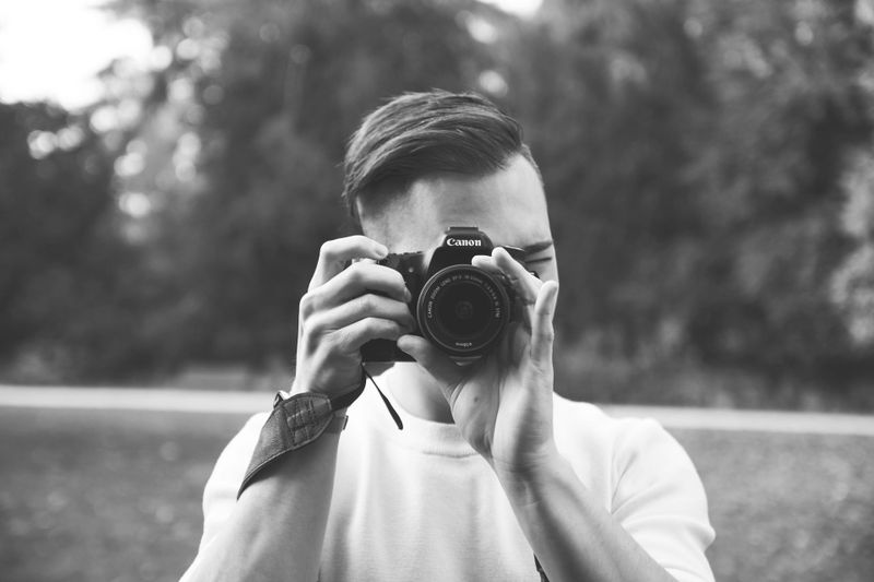 Une personne tenant un appareil photo Canon prend une photo en extérieur. Image en noir et blanc avec des arbres flous en arrière-plan.