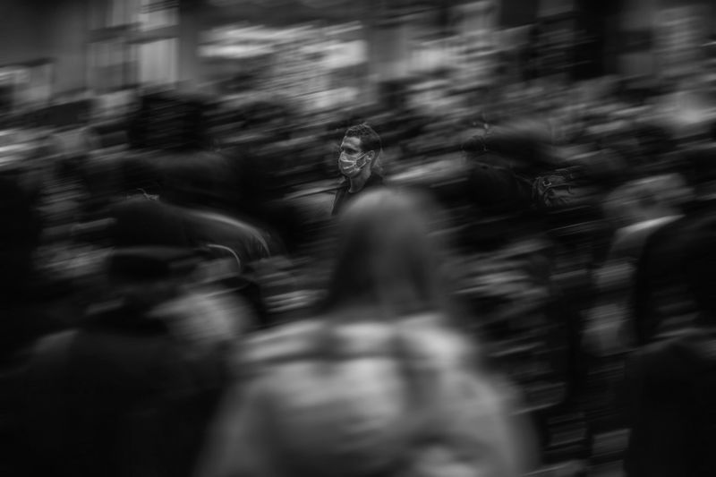 Une personne portant un masque se tient immobile au milieu d'une foule floue et animée, créant un sentiment d'isolement dans un environnement urbain animé. Noir et blanc.