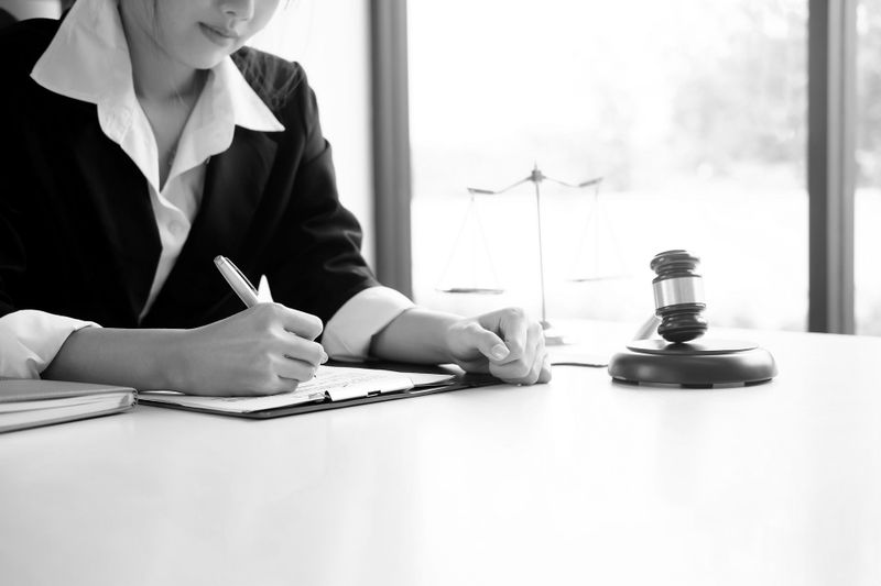 Une personne en costume écrit sur un carnet à un bureau avec un marteau et des balances de la justice à proximité ; une fenêtre lumineuse se trouve en arrière-plan.