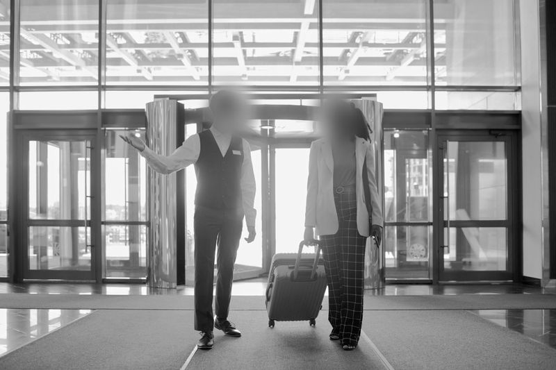 Employé d’hôtel aidant un client avec ses bagages près de l’entrée, avec des portes automatiques et un éclairage intense en arrière-plan.