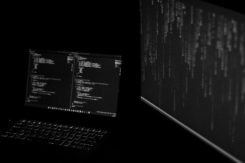 Dimly lit scene of a laptop and monitor displaying lines of code in a dark environment, creating a focused and tech-centric atmosphere.