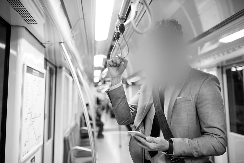 Une personne en costume se tient dans une rame de métro, tenant une barre et regardant un smartphone. L'arrière-plan montre des sièges vides et des lumières vives.