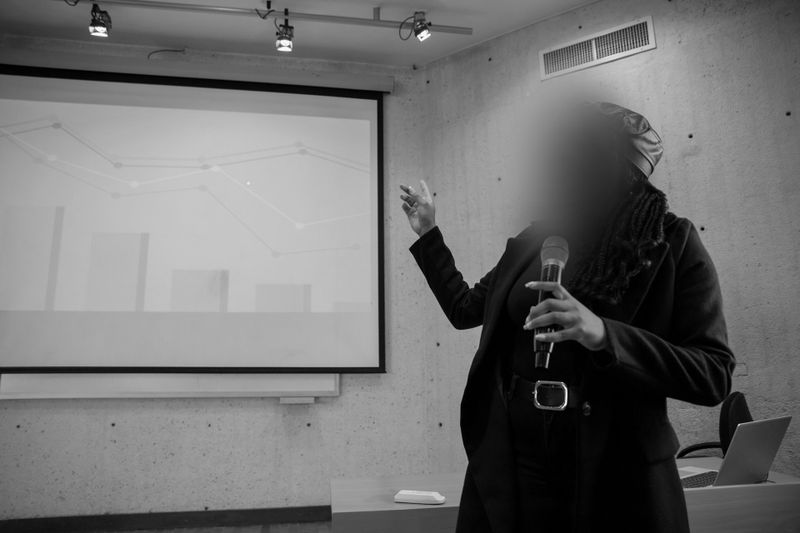 Foto en blanco y negro que muestra a una oradora, elegantemente vestida, señalando una presentación con gráficos, con el rostro desenfocado