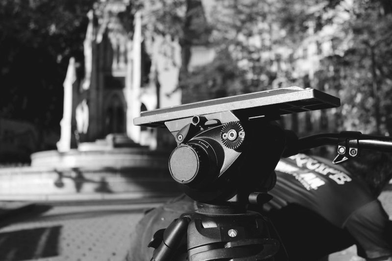 Primer plano de la cabeza de un trípode de cámara enfocado, con un fondo desenfocado de un parque y una fuente. Imagen en blanco y negro.