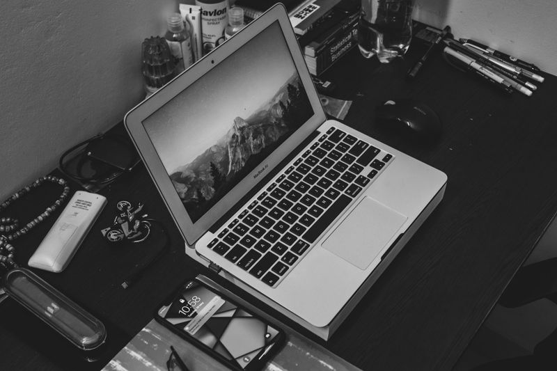 Image en noir et blanc d'un ordinateur portable affichant une scène de montagne sur un bureau encombré avec un smartphone, des stylos et divers objets.