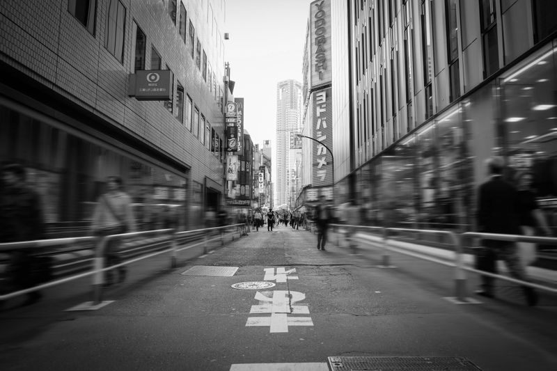 Black and white photo of a bustling city street in Japan, with tall buildings and blurred pedestrians, emphasizing movement and urban life.