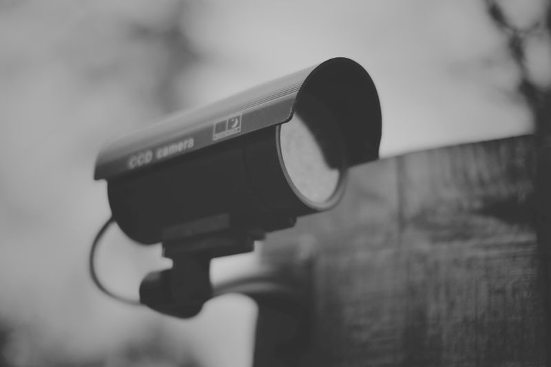 Black and white image of a security camera mounted on a wooden post, facing left.