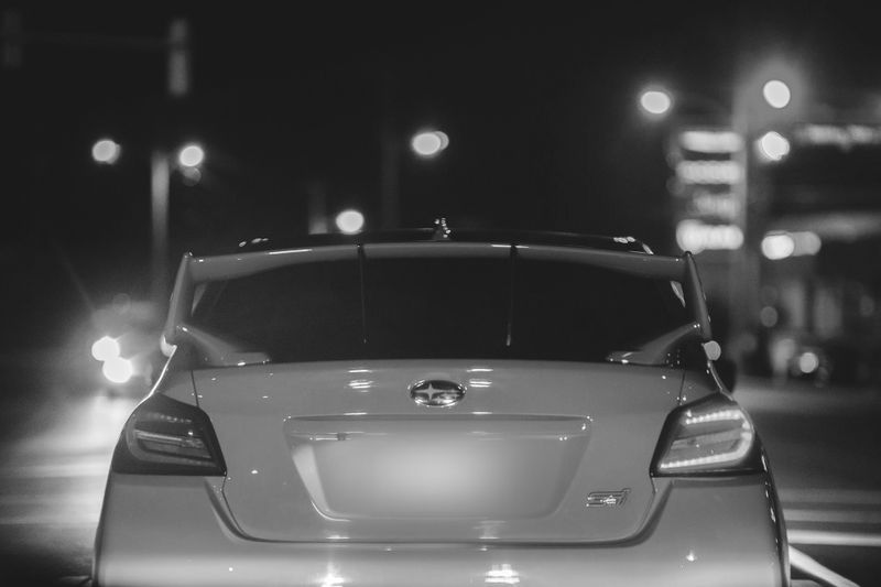 Black and white image of a car with a spoiler, seen from behind at night, with blurred city lights in the background.