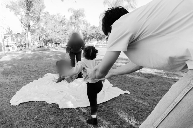 Photo en noir et blanc d’un enfant marchant sur l’herbe vers une couverture, aidé par un adulte. Une autre personne se tient sur la couverture dans un parc.