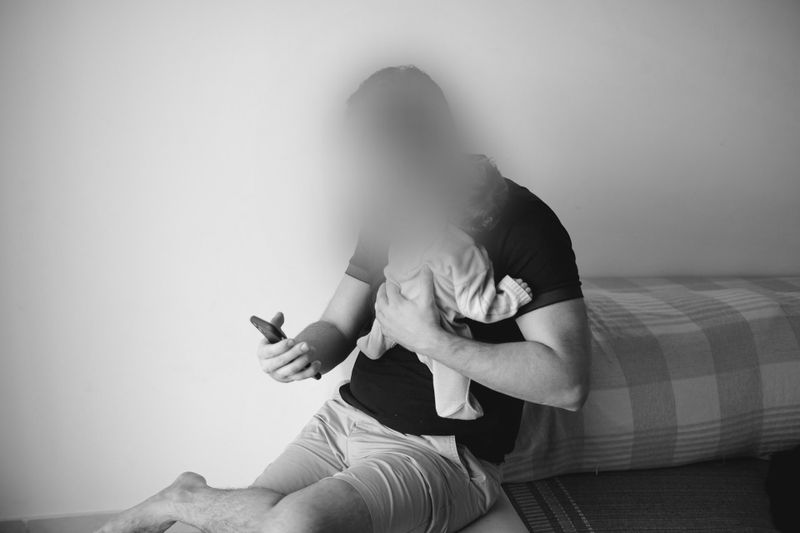 A man is sitting on the bed, holding an infant with one hand and using a smartphone with the other. The photo is black and white.