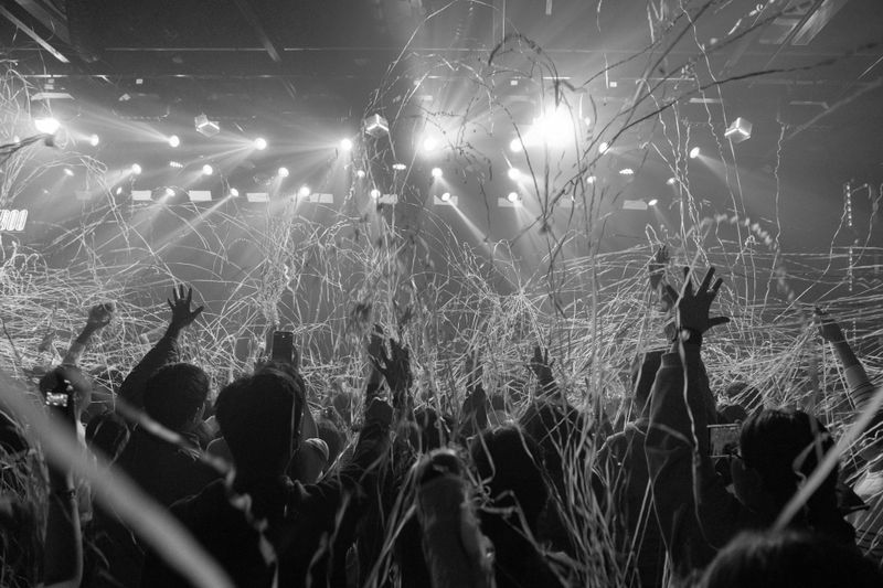 Black and white photo of a concert crowd with hands raised, surrounded by streamers and bright stage lights in the background.