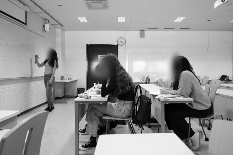 Tres estudiantes están sentados en escritorios en un aula, frente a un maestro que escribe en una pizarra blanca. El entorno es moderno y bien iluminado.