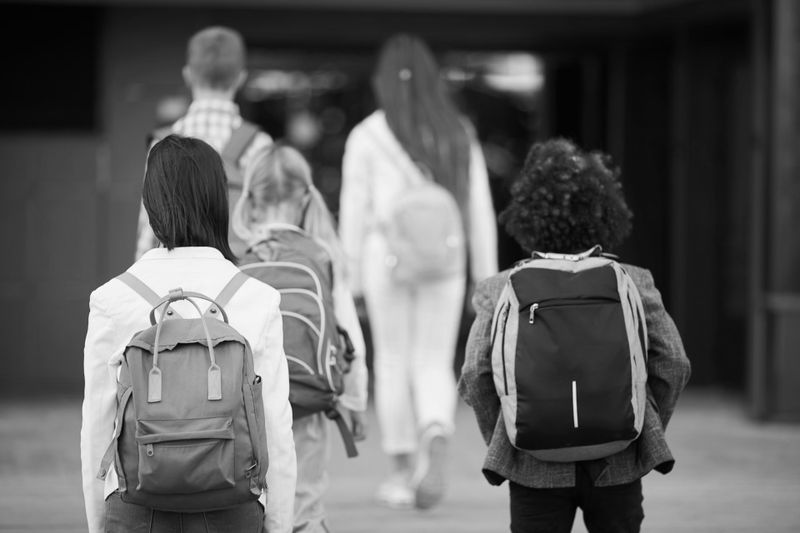 Un grupo de niños con mochilas camina hacia la entrada de un edificio, visto desde atrás, en blanco y negro.