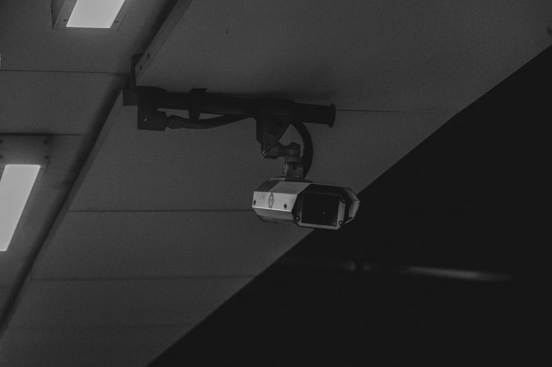 Black and white image of a security camera mounted on a ceiling, with fluorescent lights nearby.