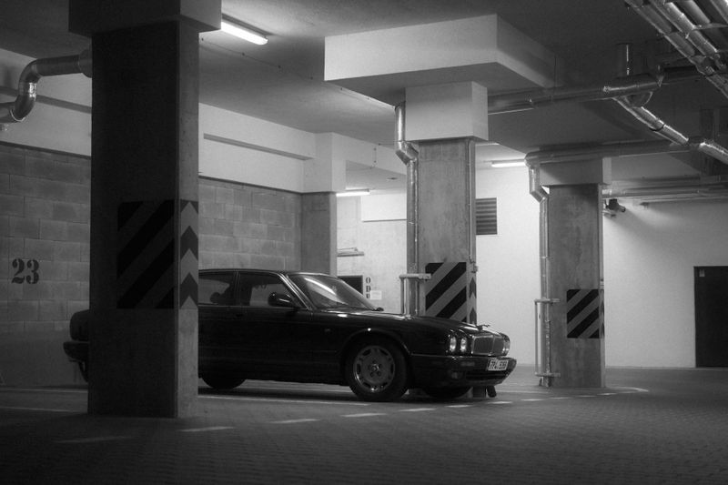 Une voiture noire est stationnée dans un parking souterrain faiblement éclairé, entourée de piliers en béton portant des marquages rayés.