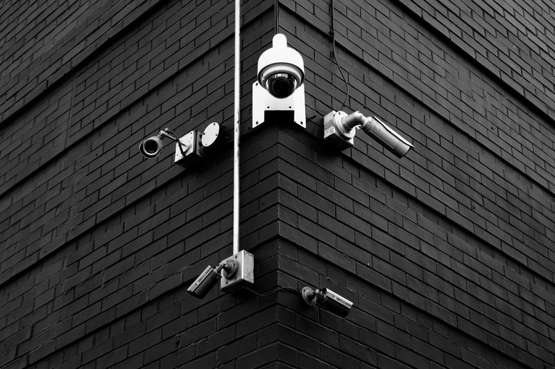 Black and white image of a building corner with multiple security cameras and pipes mounted on dark brick walls.