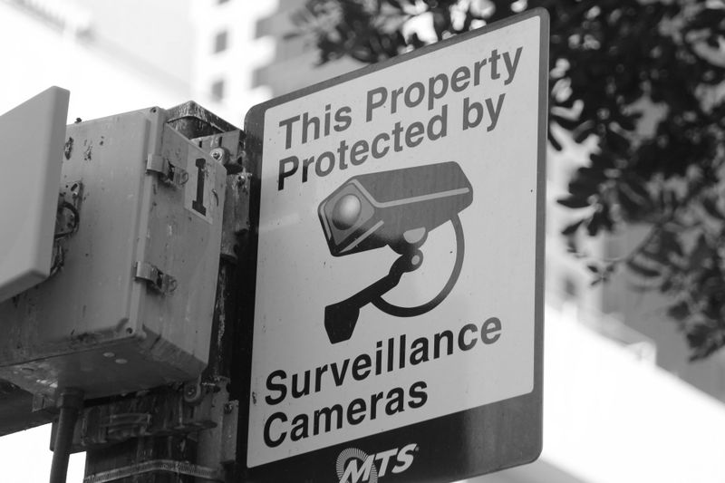 Panneau sur un poteau indiquant « Cette propriété est protégée par des caméras de surveillance » avec une image d'une caméra. Arbres et bâtiment en arrière-plan.