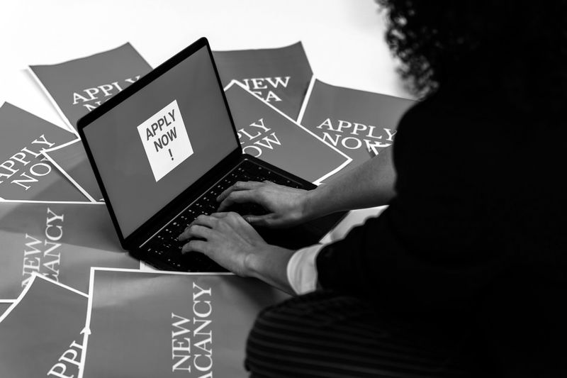 Person typing on a laptop with "Apply Now" on the screen, surrounded by papers labeled "New Vacancy."