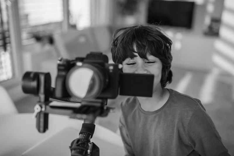 Un niño con cabello desordenado sonríe mientras mira a través de una cámara en un trípode en interiores, en una habitación con luz suave. Imagen en blanco y negro.