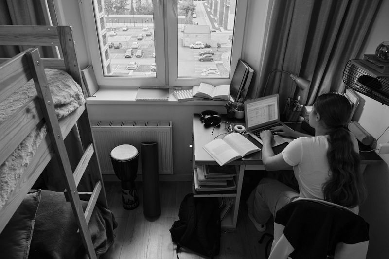 Una persona con el pelo largo trabaja en una laptop en un escritorio en una habitación ordenada con una litera, un tambor y una ventana que da a autos estacionados y edificios.