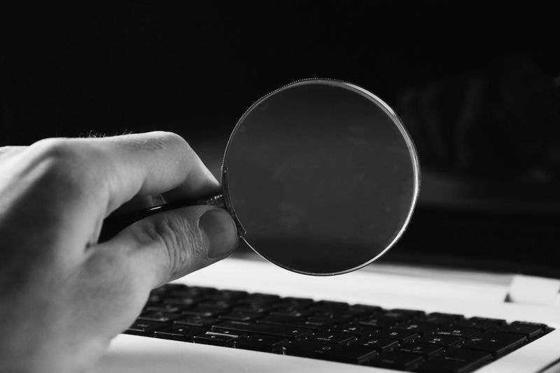 Hand holding a magnifying glass over a laptop keyboard in black and white.