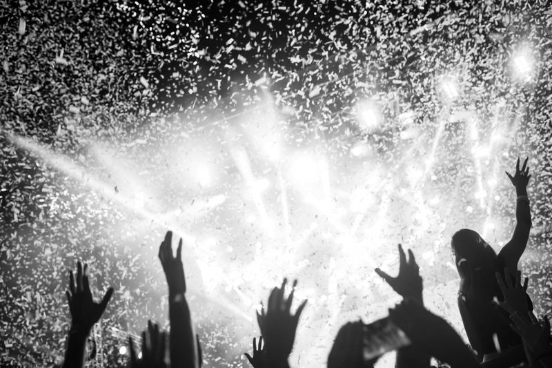 Concert crowd with raised hands, confetti falling, and bright stage lights creating a festive atmosphere. Black and white image.