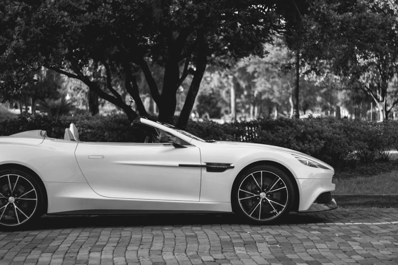 Imagen en blanco y negro de un elegante coche deportivo descapotable estacionado en una calle adoquinada, con árboles al fondo.