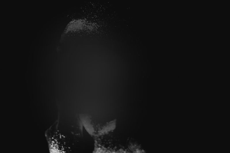Abstract dark image with a faint, obscured silhouette of a person, surrounded by scattered white particles on a black background.