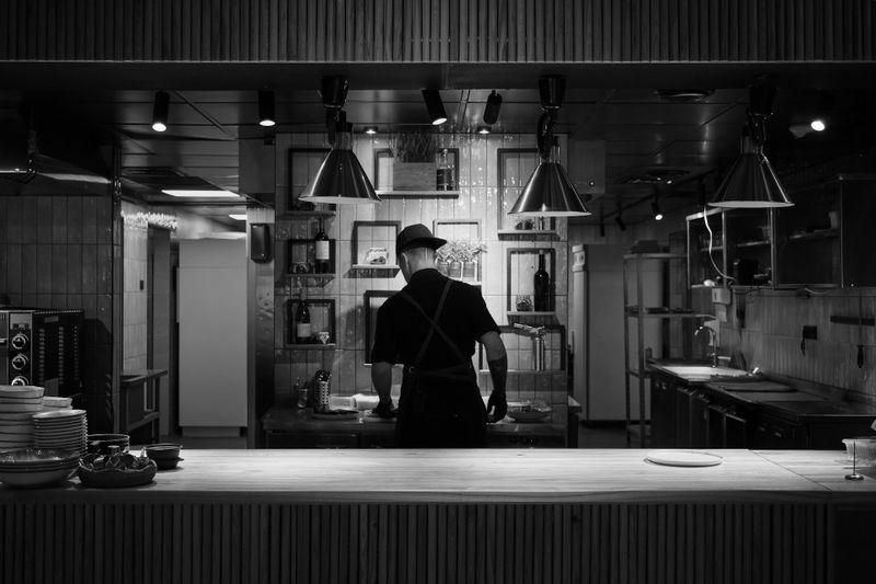 Chef trabajando solo en una cocina de estilo industrial, con poca luz, visto desde atrás, con equipo de cocina y luces colgantes visibles.