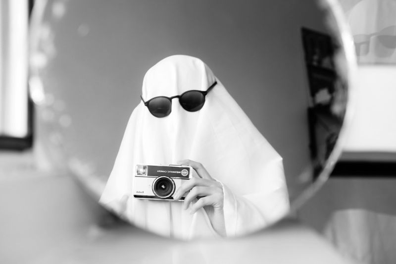 Person in a sheet ghost costume with sunglasses, holding a camera, reflected in a round mirror. Black and white image.