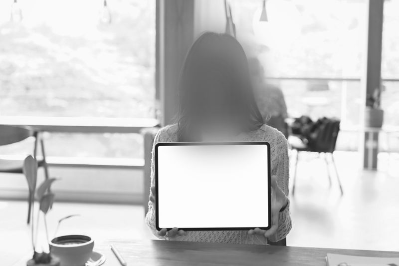 Person holding a blank tablet screen in a cafe setting, with a blurred background and a coffee cup on the table in the foreground.