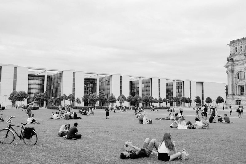 Personas relajándose en un césped con edificios modernos y parte de una estructura histórica al fondo, bicicletas estacionadas cerca.