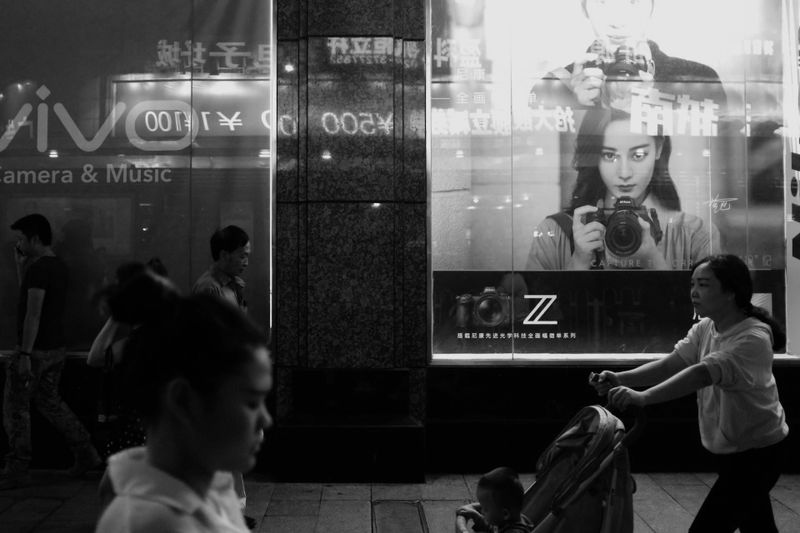 Photo en noir et blanc de personnes passant devant une grande publicité pour appareil photo dans une rue de la ville la nuit.