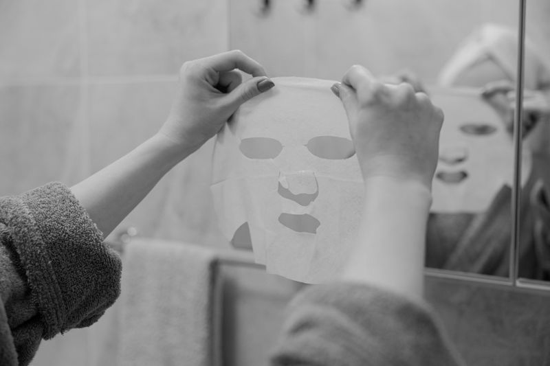 Person holding a sheet mask in front of a bathroom mirror, wearing a towel and robe.