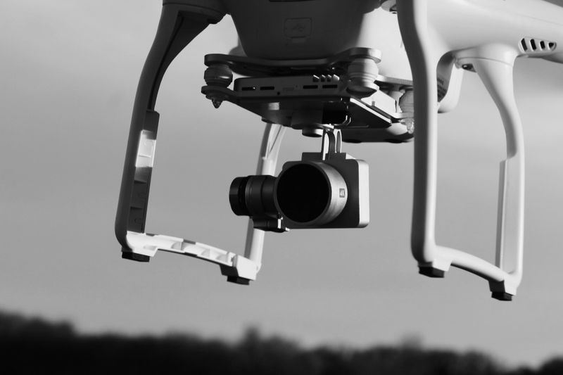 Close-up of a drone with a mounted camera, flying against a blurred sky and landscape in black and white.