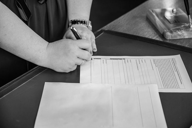 Una persona firmando un documento en un escritorio, con un reloj en la muñeca. La imagen está en blanco y negro.