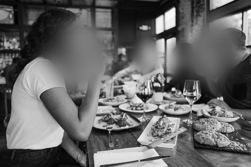 Personas sentadas en una mesa de restaurante disfrutando de una comida con copas de vino y varios platos, en un ambiente acogedor y con poca luz. Los rostros están borrosos.
