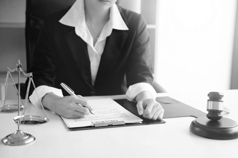 Une personne en costume écrit sur un document à un bureau, avec une balance de la justice et un marteau à proximité.