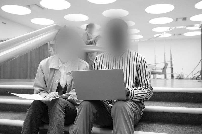 Deux personnes aux visages flous sont assises sur des marches, utilisant un ordinateur portable. En arrière-plan, des plafonniers circulaires et un intérieur moderne.