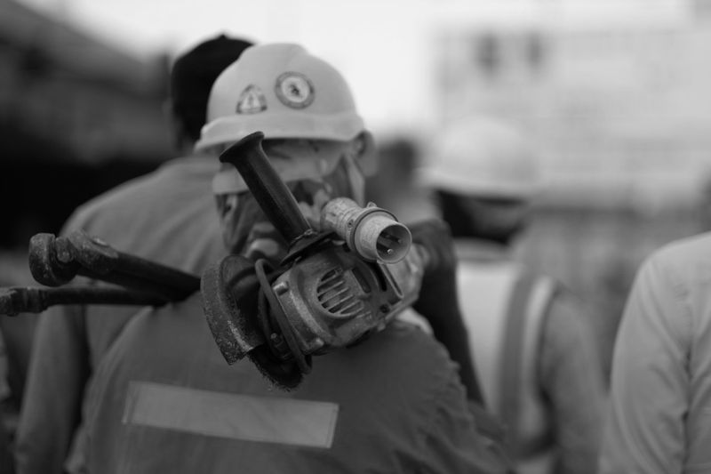 Un trabajador de la construcción con casco lleva una herramienta eléctrica sobre el hombro en un sitio de trabajo, con otros trabajadores desenfocados en el fondo.