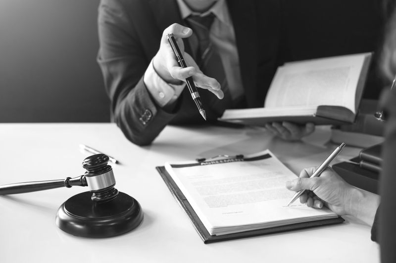 Deux personnes en discussion à un bureau avec des documents, un marteau et un stylo. Une personne fait un geste tandis que l'autre écrit. Image en noir et blanc.