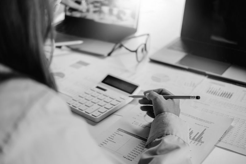 Una persona usando una calculadora mientras revisa documentos financieros en un escritorio con laptops y gafas.