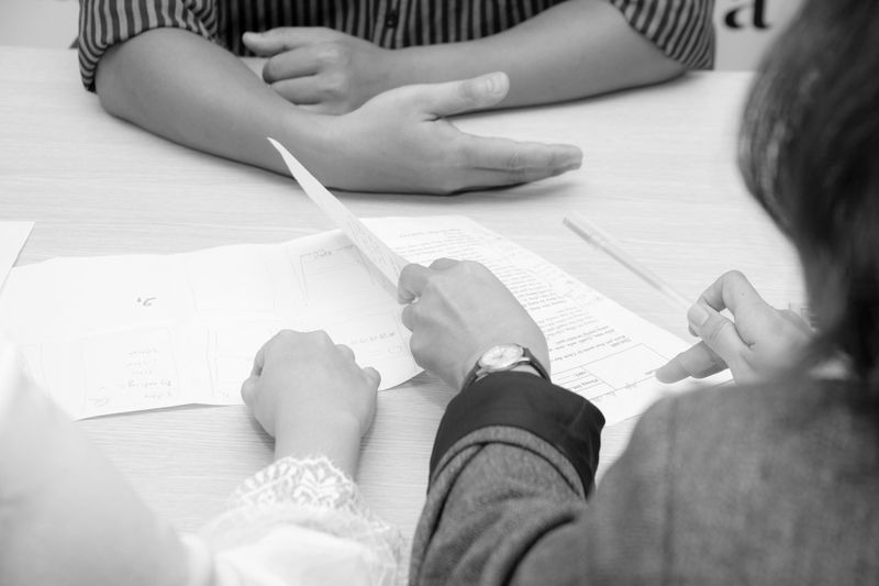 Image en noir et blanc de trois personnes assises à une table, en train de discuter de documents. Les mains et les papiers sont visibles.