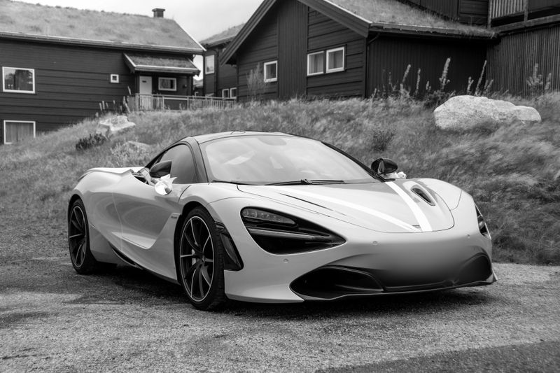 En la foto en blanco y negro se ve un coche brillante de la marca McLaren, modelo 720S, frente a casas noruegas