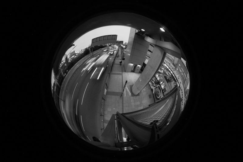 Fisheye lens view of an urban street with light trails from passing cars and pedestrians walking on the sidewalk at dusk.