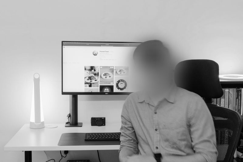 Une personne au visage flou est assise à un bureau avec un ordinateur affichant un site Web sur la nourriture, à côté d'une lampe moderne et d'un appareil photo.